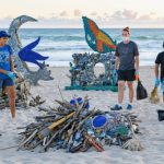 A multi-generational group of community members engaged in a beach cleanup at sunrise, with art installations made from marine debris in the background, showcasing the integration of traditional maritime culture and modern environmental stewardship.