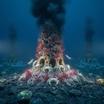 Black smoker vent chimney emitting a dark plume on the deep seafloor, surrounded by giant red-plumed tube worms, pale crabs, and eyeless shrimp, lit by submersible lights with distant chimneys and rocky seabed fading into darkness