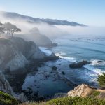 Wide view from a seaside bluff of marine layer fog rolling over the California coastline, veiling rocky cliffs and tide pools with wind-bent cypress and distant headlands in soft early morning light