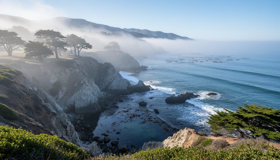 Wide view from a seaside bluff of marine layer fog rolling over the California coastline, veiling rocky cliffs and tide pools with wind-bent cypress and distant headlands in soft early morning light