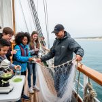 Students and a marine educator on a two-masted schooner collect a plankton tow and inspect a water sample at a microscope on Lake Michigan, with blue water and distant sand dunes in the background.