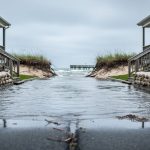 Low-angle view of a flooded coastal street with seawater over the curb, sandbags at doorways, and modest seaside homes under an overcast sky, with dunes and marsh grasses in the softly blurred background.