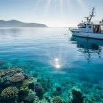 Sunlit coral reef beneath clear coastal water with a small anchored research vessel in the foreground, representing a marine protected area.