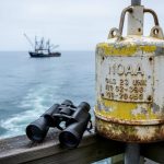 Research buoy and coastal monitoring equipment on a dock with ocean waves and a distant fishing vessel under daylight.