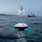 A sensor-equipped research buoy on the ocean surface at dusk, with a distant research vessel and floating debris suggesting advanced pollution detection monitoring.