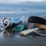Close-up of wet shoreline debris showing plastic pieces, metal can fragments, glass shards, rubber, textiles, processed wood splinters, and paper or cardboard in sand.