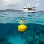 “A research vessel anchored above clear blue-green water with a floating buoy indicating a marine protected zone near a reef.”