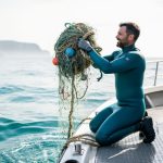Volunteer on a small boat removing tangled ghost fishing gear from the ocean, with clear blue water and distant shoreline in the background.