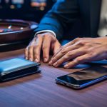 “Hands resting on a casino poker table beside a closed wallet, a face-down smartphone, and a wristwatch, suggesting gambling safety boundaries and secure access.”