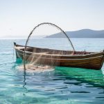 A wooden fishing boat near a clear turquoise coast with nets being hauled, representing sustainable fisheries and ocean protection.